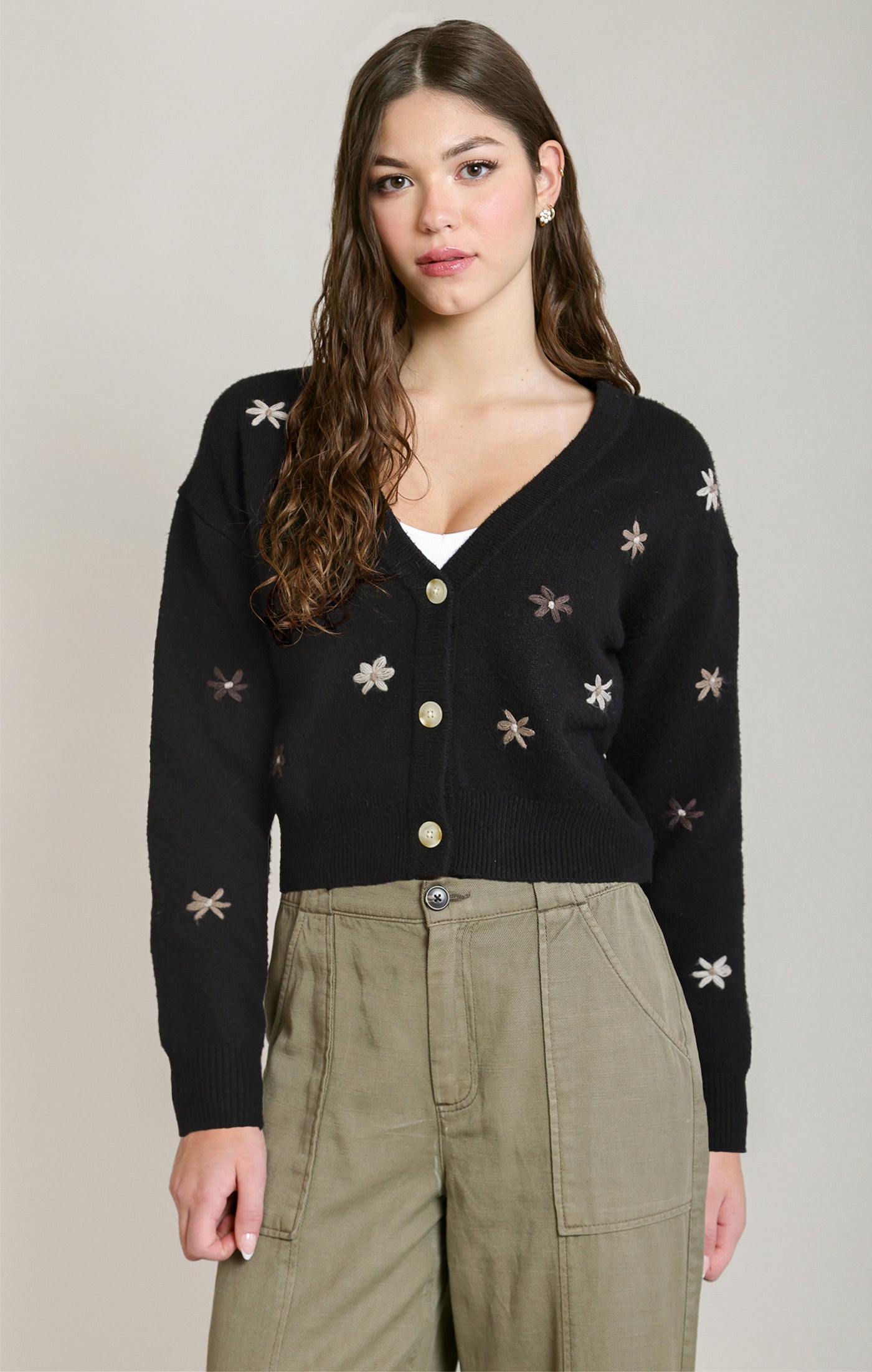 A woman modeling a black knit cardigan with embroidered star and snowflake patterns, featuring gold buttons and a V-neckline, paired with olive green cargo pants against a light gray studio background.