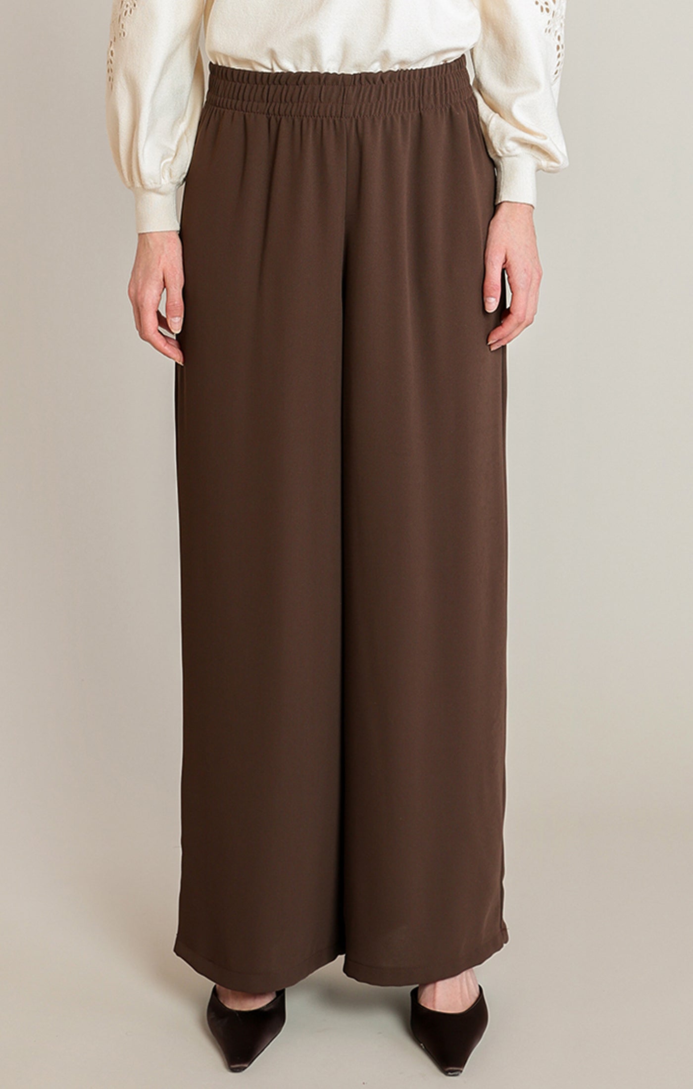 A model is wearing a pair of wide-leg brown pants with an elastic waistband, styled with a cream blouse and black pointed-toe heels against a neutral studio background.
